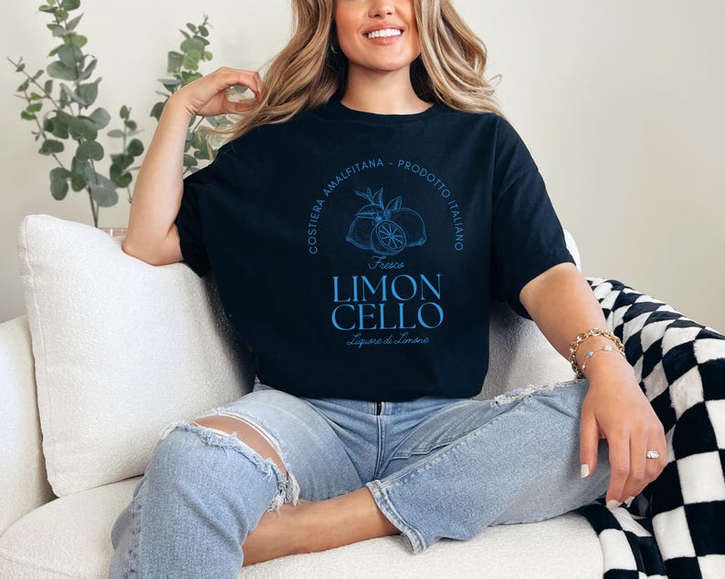 Black T-shirt with a vibrant blue design reading Costiera Amalfitana - Prodotto Italiano - Limoncello. Minimalist beverage label style. Worn by a woman in jeans, sitting casually. Perfect for limoncello lovers and Italian culture fans.