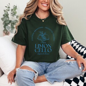 Forest green T-shirt with a vibrant blue design reading Costiera Amalfitana - Prodotto Italiano - Limoncello. Minimalist beverage label style. Worn by a woman in jeans, sitting casually. Perfect for limoncello lovers and Italian culture fans.