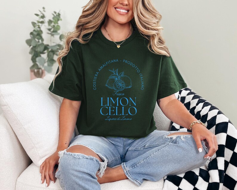Forest green T-shirt with a vibrant blue design reading Costiera Amalfitana - Prodotto Italiano - Limoncello. Minimalist beverage label style. Worn by a woman in jeans, sitting casually. Perfect for limoncello lovers and Italian culture fans.
