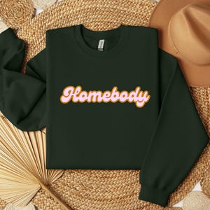 Folded forest green sweatshirt with a retro cursive design reading Homebody in bold pink with a coffee brown outline. Showcasing the cozy quality of the fabric. Perfect for those who love the comfort of home.