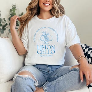 White T-shirt with a vibrant blue design reading Costiera Amalfitana - Prodotto Italiano - Limoncello. Minimalist beverage label style. Worn by a woman in jeans, sitting casually. Perfect for limoncello lovers and Italian culture fans.