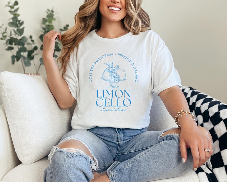 White T-shirt with a vibrant blue design reading Costiera Amalfitana - Prodotto Italiano - Limoncello. Minimalist beverage label style. Worn by a woman in jeans, sitting casually. Perfect for limoncello lovers and Italian culture fans.