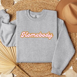 Folded sport grey sweatshirt with a retro cursive design reading Homebody in bold pink with a coffee brown outline. Showcasing the cozy quality of the fabric. Perfect for those who love the comfort of home.