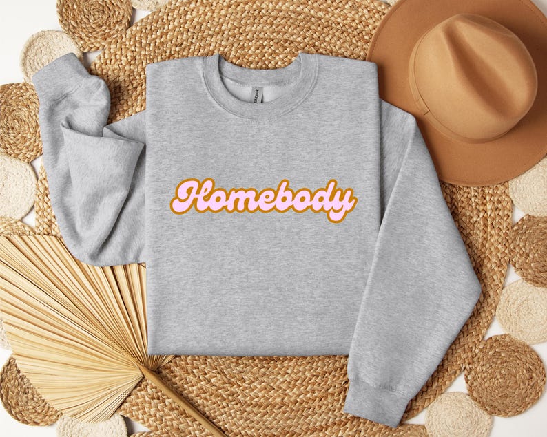 Folded sport grey sweatshirt with a retro cursive design reading Homebody in bold pink with a coffee brown outline. Showcasing the cozy quality of the fabric. Perfect for those who love the comfort of home.