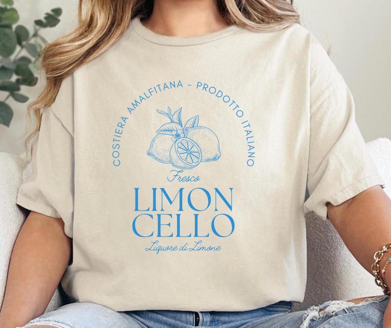 Sand T-shirt with a vibrant blue design reading Costiera Amalfitana - Prodotto Italiano - Limoncello. Minimalist beverage label style. Worn by a woman in jeans, sitting casually. Perfect for limoncello lovers and Italian culture fans.