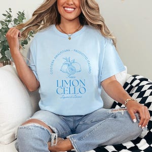 Light blue T-shirt with a vibrant blue design reading Costiera Amalfitana - Prodotto Italiano - Limoncello. Minimalist beverage label style. Worn by a woman in jeans, sitting casually. Perfect for limoncello lovers and Italian culture fans.