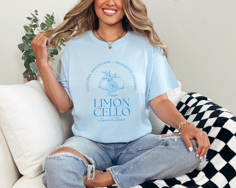 Light blue T-shirt with a vibrant blue design reading Costiera Amalfitana - Prodotto Italiano - Limoncello. Minimalist beverage label style. Worn by a woman in jeans, sitting casually. Perfect for limoncello lovers and Italian culture fans.
