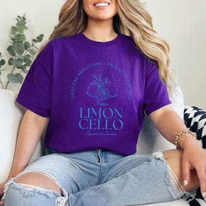 Purple T-shirt with a vibrant blue design reading Costiera Amalfitana - Prodotto Italiano - Limoncello. Minimalist beverage label style. Worn by a woman in jeans, sitting casually. Perfect for limoncello lovers and Italian culture fans.