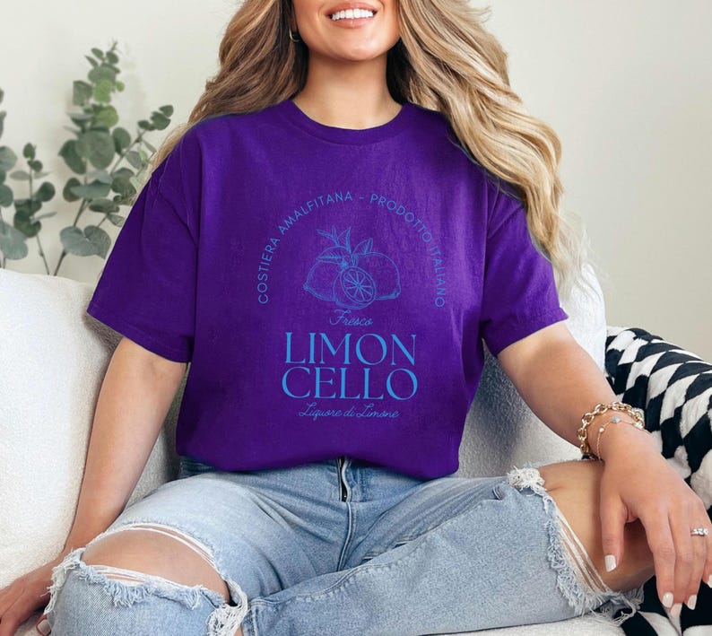 Purple T-shirt with a vibrant blue design reading Costiera Amalfitana - Prodotto Italiano - Limoncello. Minimalist beverage label style. Worn by a woman in jeans, sitting casually. Perfect for limoncello lovers and Italian culture fans.