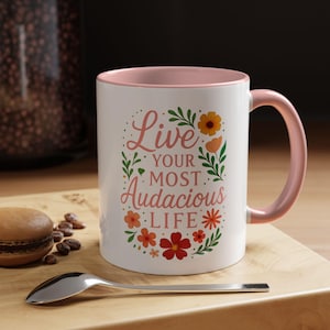 Puede incluir: Taza de cerámica blanca con interior y asa rosas. La taza presenta el texto "Live Your Most Audacious Life" rodeado de ilustraciones florales en naranja, rojo y verde. Una cuchara y un pastelito están sobre una superficie de madera.