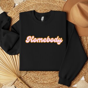 Folded black sweatshirt with a retro cursive design reading Homebody in bold pink with a coffee brown outline. Showcasing the cozy quality of the fabric. Perfect for those who love the comfort of home.