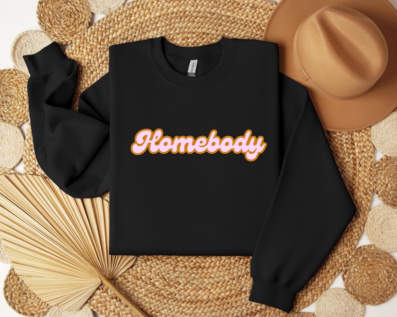 Folded black sweatshirt with a retro cursive design reading Homebody in bold pink with a coffee brown outline. Showcasing the cozy quality of the fabric. Perfect for those who love the comfort of home.