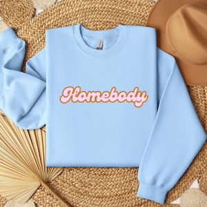 Folded light blue sweatshirt with a retro cursive design reading Homebody in bold pink with a coffee brown outline. Showcasing the cozy quality of the fabric. Perfect for those who love the comfort of home.