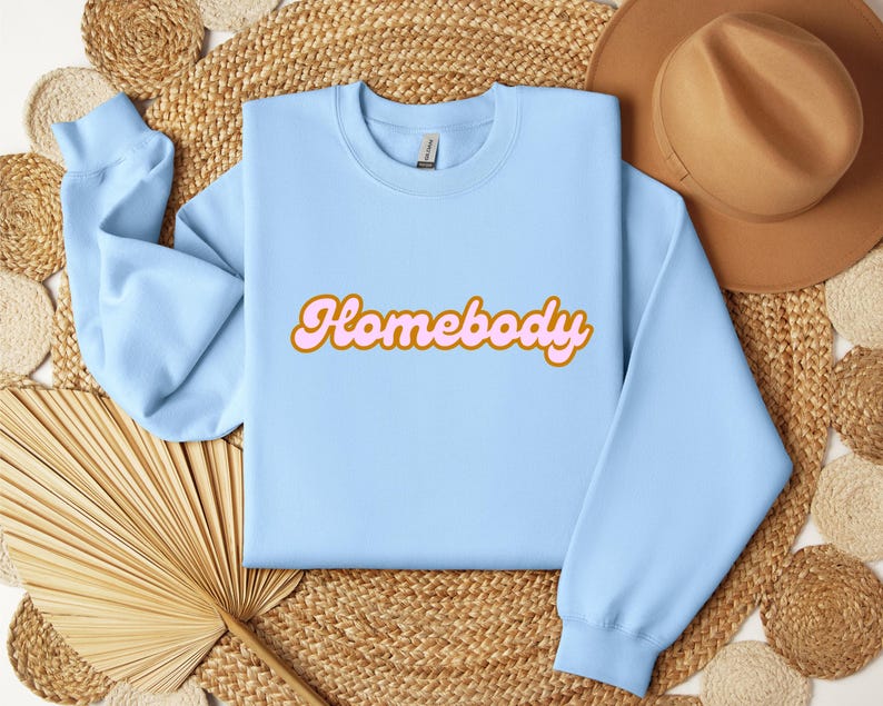 Folded light blue sweatshirt with a retro cursive design reading Homebody in bold pink with a coffee brown outline. Showcasing the cozy quality of the fabric. Perfect for those who love the comfort of home.