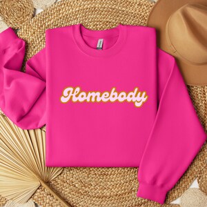 Folded heliconia pink sweatshirt with a retro cursive design reading Homebody in bold pink with a coffee brown outline. Showcasing the cozy quality of the fabric. Perfect for those who love the comfort of home.