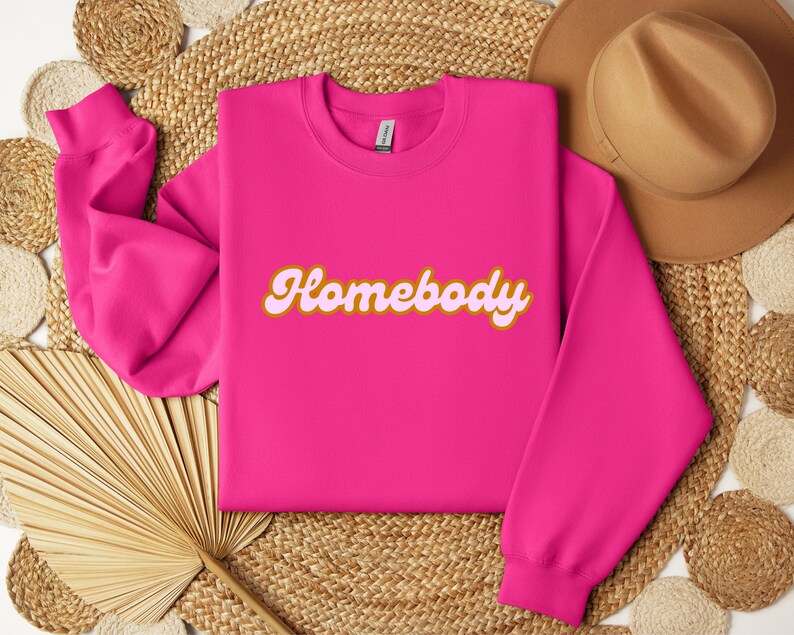 Folded heliconia pink sweatshirt with a retro cursive design reading Homebody in bold pink with a coffee brown outline. Showcasing the cozy quality of the fabric. Perfect for those who love the comfort of home.