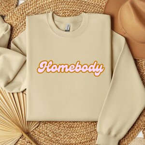 Folded sand sweatshirt with a retro cursive design reading Homebody in bold pink with a coffee brown outline. Showcasing the cozy quality of the fabric. Perfect for those who love the comfort of home.