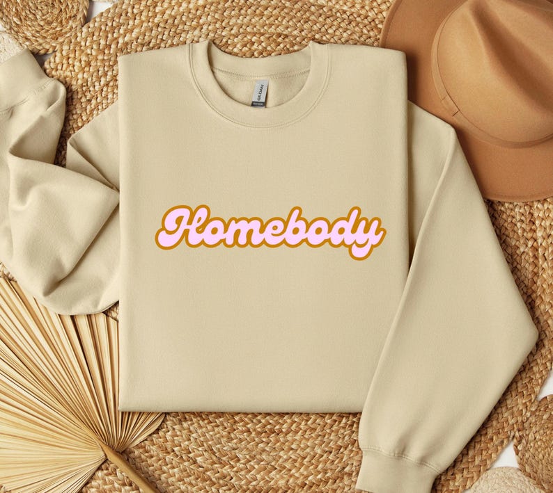 Folded sand sweatshirt with a retro cursive design reading Homebody in bold pink with a coffee brown outline. Showcasing the cozy quality of the fabric. Perfect for those who love the comfort of home.