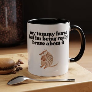 May include: White ceramic mug with a black interior and handle. The mug features the text "my tummy hurts but im being really brave about it" and an illustration of an armadillo. A spoon and a macaron are in the foreground.
