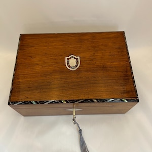 May include: A rectangular wooden box with a polished brown finish, featuring a silver-toned shield-shaped inlay on the lid. The edges are decorated with dark, inlaid geometric patterns and mother-of-pearl accents. A silver key with a gray tassel is inserted.