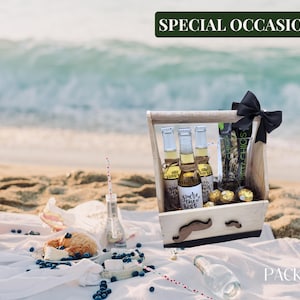 Puede incluir: Una caja de regalo de madera con un lazo negro y un dise&ntilde;o de bigote. La caja contiene tres botellas de cerveza, una bolsa de aperitivos y caramelos de chocolate. La caja est&aacute; sentada sobre una manta blanca con ar&aacute;ndanos y un pastel.