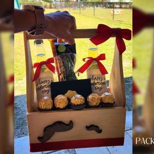 Puede incluir: Una caja de regalo de madera con una cinta roja y dos botellas de cerveza. Las botellas est&aacute;n decoradas con el texto "Happy Valentine's Day". La caja tambi&eacute;n contiene una peque&ntilde;a pizarra negra y caramelos de chocolate.