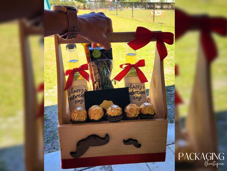 Puede incluir: Una caja de regalo de madera con una cinta roja y dos botellas de cerveza. Las botellas est&aacute;n decoradas con el texto "Happy Valentine's Day". La caja tambi&eacute;n contiene una peque&ntilde;a pizarra negra y caramelos de chocolate.