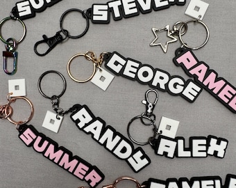 May include: A collection of colorful keychains with different names printed on them, including "Summer", "Steven", "George", "Randy", "Alex", and "Pamela". Each keychain has a small square charm attached to it.