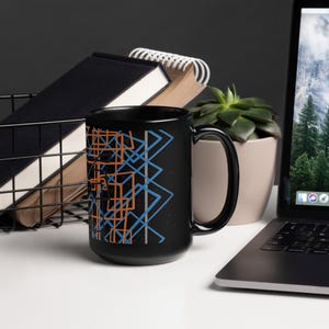 May include: Black ceramic mug with a geometric design in orange, blue, and white. The design features overlapping squares and triangles.