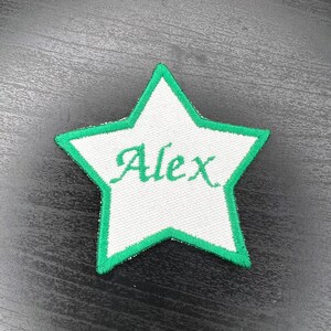 May include: A white star-shaped patch with a green border and the name "Alex" embroidered in green script. The patch is likely made of fabric and designed for application on clothing or accessories.