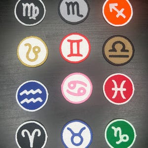 May include: A collection of twelve round embroidered patches, each featuring a different zodiac symbol. The patches are in various colors, including black, gray, orange, white, red, brown, blue, pink, and green, with white stitching.