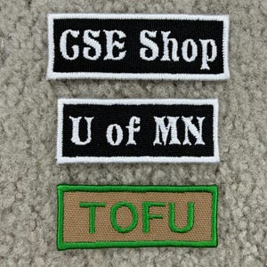 K&ouml;nnte beinhalten: Drei rechteckige, gestickte Aufn&auml;her. Der obere Aufn&auml;her ist schwarz mit wei&szlig;em Text "GSE Shop". Der mittlere Aufn&auml;her ist schwarz mit wei&szlig;em Text "U of MN". Der untere Aufn&auml;her ist beige mit gr&uuml;nem Text "TOFU".