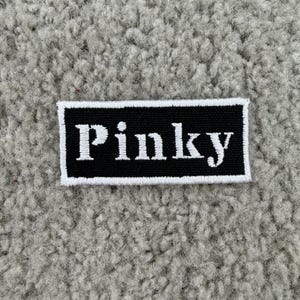 K&ouml;nnte beinhalten: Ein rechteckiger schwarzer Aufn&auml;her mit dem Wort "Pinky" in Wei&szlig; gestickt, umrahmt von einem wei&szlig;en Rand. Der Aufn&auml;her befindet sich auf einem strukturierten, hellgrauen Hintergrund.