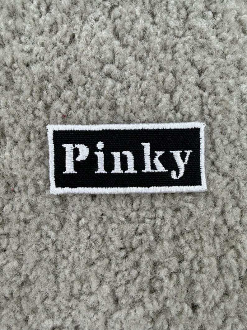 K&ouml;nnte beinhalten: Ein rechteckiger schwarzer Aufn&auml;her mit dem Wort "Pinky" in Wei&szlig; gestickt, umrahmt von einem wei&szlig;en Rand. Der Aufn&auml;her befindet sich auf einem strukturierten, hellgrauen Hintergrund.