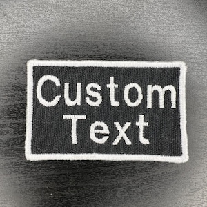 May include: A rectangular black patch with white embroidered text that reads "Custom Text." The patch has a white border and is likely designed for customization.