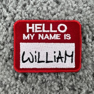 May include: A red name tag patch with white embroidered text that reads "HELLO MY NAME IS" above the name "WILLIAM" in black. The patch has a white rectangular section for the name.