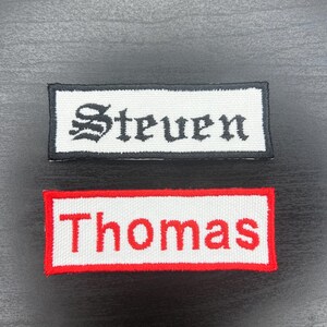 K&ouml;nnte beinhalten: Zwei rechteckige Namensschilder. Eines hat einen wei&szlig;en Hintergrund mit dem Namen "Steven" in schwarzer gotischer Schrift, umrahmt von einem schwarzen Rand. Das andere hat einen wei&szlig;en Hintergrund mit dem Namen "Thomas" in roter Schrift, umrahmt von einem roten Rand.