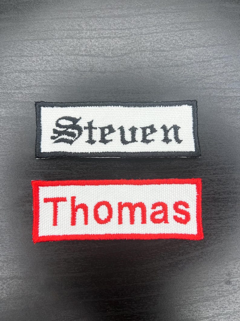 K&ouml;nnte beinhalten: Zwei rechteckige Namensschilder. Eines hat einen wei&szlig;en Hintergrund mit dem Namen "Steven" in schwarzer gotischer Schrift, umrahmt von einem schwarzen Rand. Das andere hat einen wei&szlig;en Hintergrund mit dem Namen "Thomas" in roter Schrift, umrahmt von einem roten Rand.