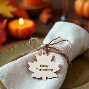 May include: Thanksgiving table setting with a cream-colored napkin, a wooden leaf-shaped tag that says "Happy Thanksgiving", and a twine bow. The plate is set on a brown charger, with pumpkins and candles in the background.