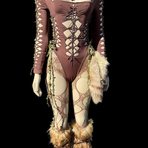 May include: A brown bodysuit with cut-out details and long sleeves, adorned with faux fur accents and decorative vines. The outfit includes matching faux fur leg warmers and a pendant necklace. The mannequin is set against a black background.