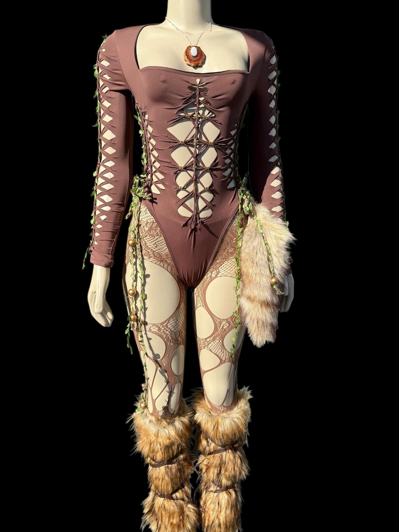 May include: A brown bodysuit with cut-out details and long sleeves, adorned with faux fur accents and decorative vines. The outfit includes matching faux fur leg warmers and a pendant necklace. The mannequin is set against a black background.