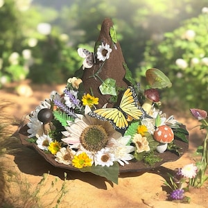 Puede incluir: Un sombrero de bruja marrón decorado con una gran mariposa amarilla, girasoles, margaritas y setas. El sombrero tiene un ala ancha y una corona alta y puntiaguda. El sombrero está adornado con varias flores y vegetación.