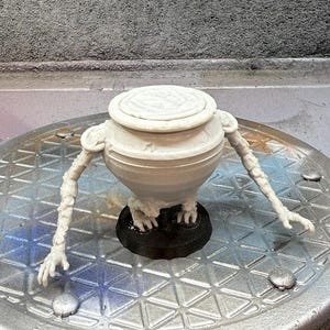 May include: A white, 3D-printed fantasy creature with a pot-like body, arms, and hands. The creature stands on a black base. The arms are extended, and the hands are open. The background is a textured grey surface.