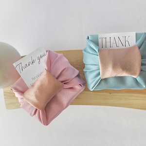 May include: Two square fabric-wrapped gifts, one in dusty pink and one in light blue, each featuring a metallic rose gold band across the center. Each package includes a white card with black text reading Thank you and THANK.