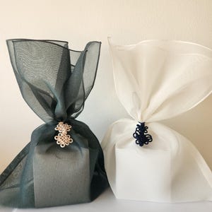 May include: Two gift bags made of sheer organza fabric, one in dark green and one in white. Each bag is gathered at the top and secured with a decorative knotted cord ornament. The dark green bag features a cream-colored knot, while the white bag features a dark blue knot.