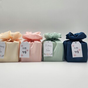 May include: Four wrapped gift boxes in pastel colors: cream, pink, mint green, and navy blue. Each box is tied with a matching fabric and a tag. The tags have Korean text and floral accents.