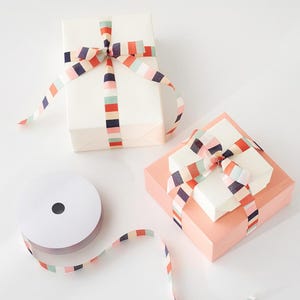 Puede incluir: Dos regalos envueltos con cinta a rayas de colores y un rollo de cinta a juego. Un regalo es blanco, el otro es melocotón. La cinta presenta rayas rojas, azules y melocotón.