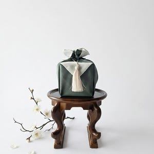May include: A dark green fabric gift wrap with white triangular accents and a cream-colored tassel, tied in a knot at the top. The package sits on a small, dark brown wooden pedestal table next to a delicate branch with white blossoms.
