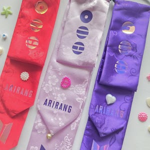 Può includere: Tre accessori per capelli in rosso, viola chiaro e viola, ciascuno dotato di un elastico nero, una coda in tessuto con motivo floreale, tre accenti olografici circolari, una piccola perlina decorativa, il testo ARIRANG e un logo sul fondo.