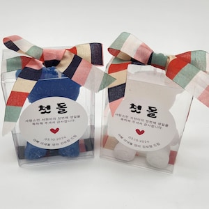 May include: Two bear-shaped candles, one blue and one white, each inside a clear plastic box topped with a multi-colored striped ribbon. A circular white label on each box features Korean text, a red heart icon, and the date 03.10.2024.